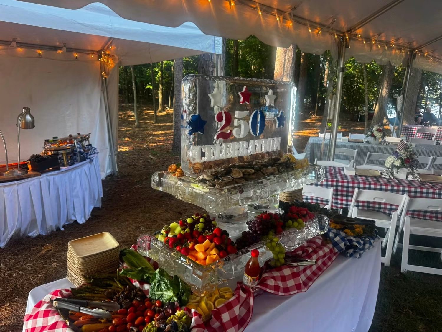A carved ice centrepiece over a raw bar of oysters and a fruit and vegetable spread, under a marquee strung with lights