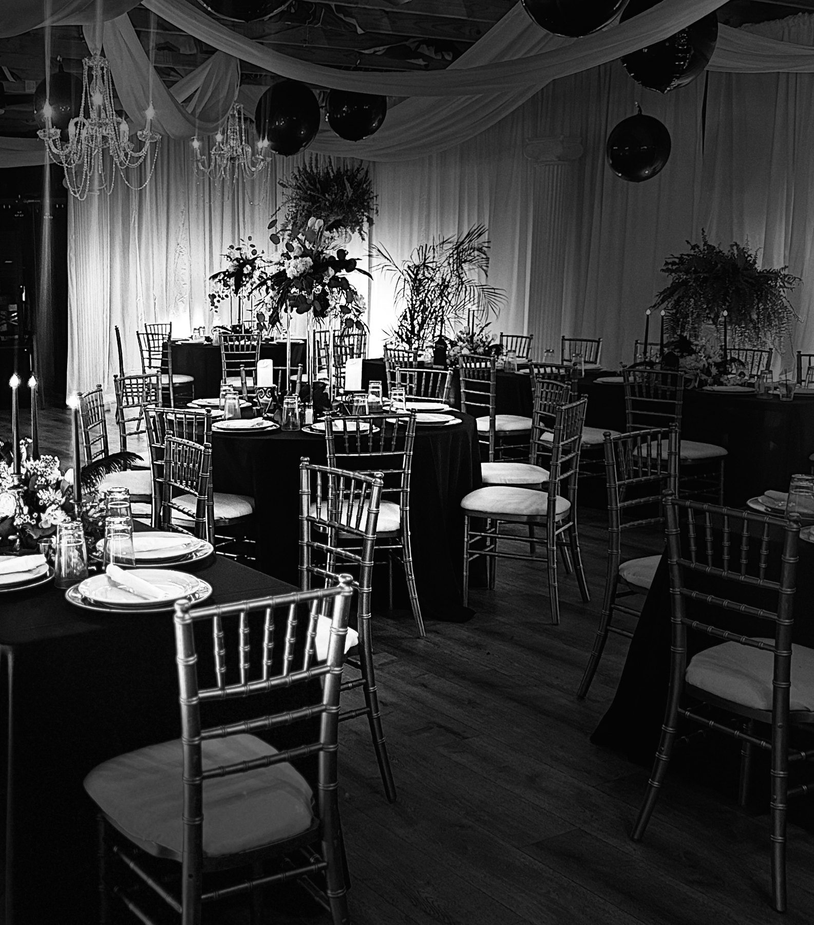 A ballroom set for a black-tie dinner: ceiling draping, chandeliers, gold chairs, tall florals and black linen
